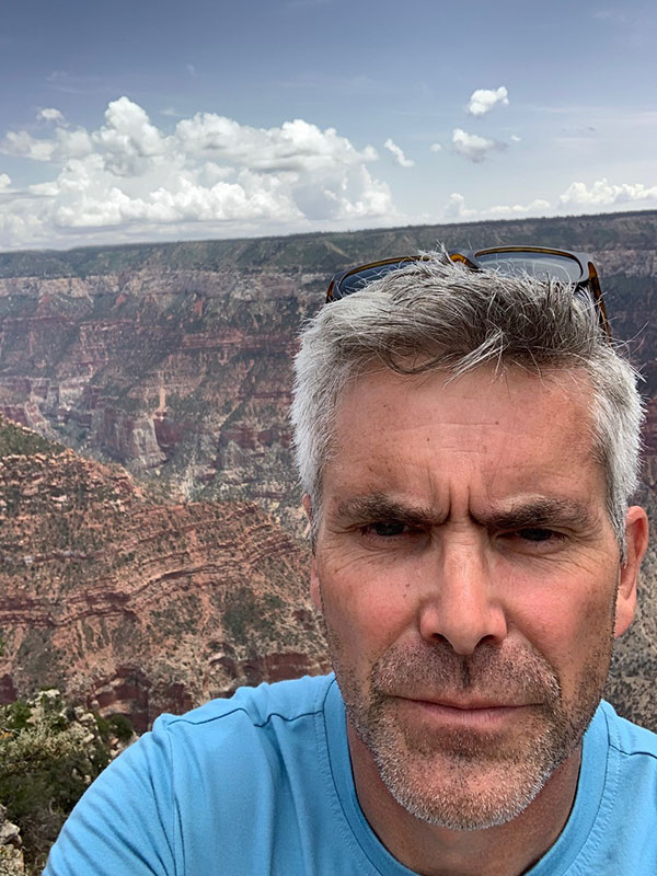 A headshot of Rich Hardy, who poses outdoors in a blue shirt, with the grand canyon behind him.