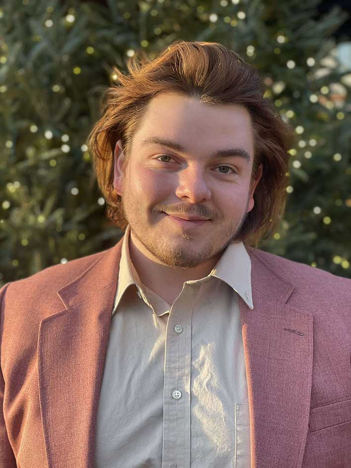 A headshot of Wyatt Reed, who wears a light red blazer and poses outdoors.